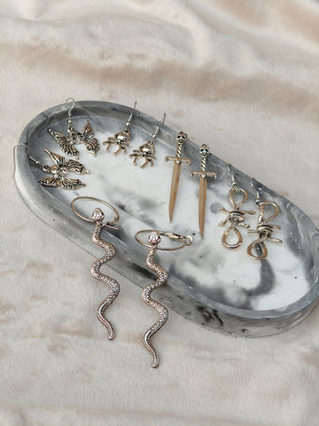 Midnight Grunge 5-Pair Silver Gothic Earring Set on a marble tray, featuring textured snake hoops, sword dangles, skull butterflies, spiders, and witch knot earrings by Marjan Collections